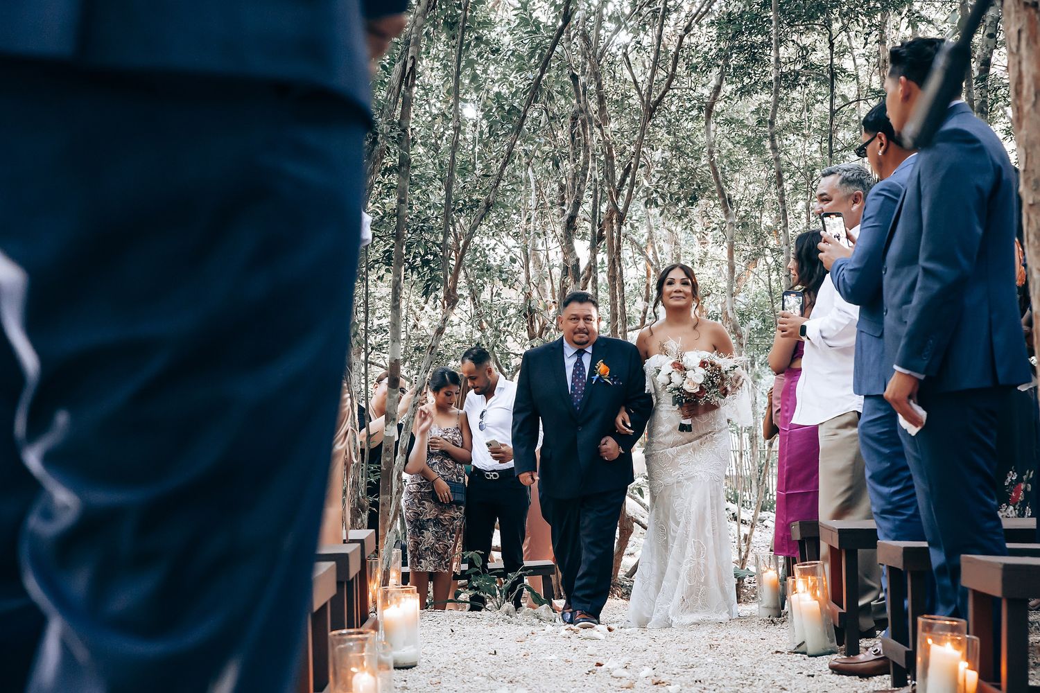 Wedding party gathers in a wooded outdoor ceremony location with guests seated on rustic wooden benches.