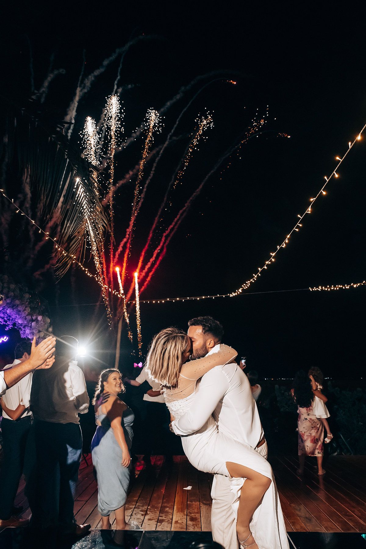 Celebration on outdoor dance floor with fireworks and string lights illuminating the night sky.