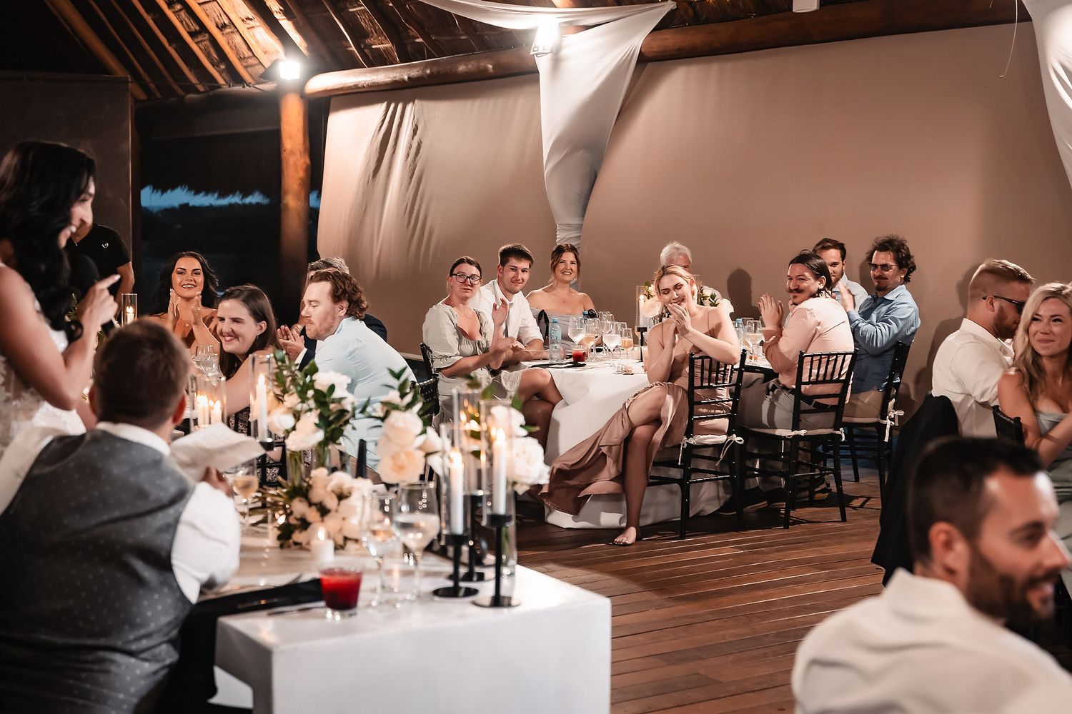 Wedding reception guests seated at long tables in a barn venue with white ceiling drapes and ambient lighting.