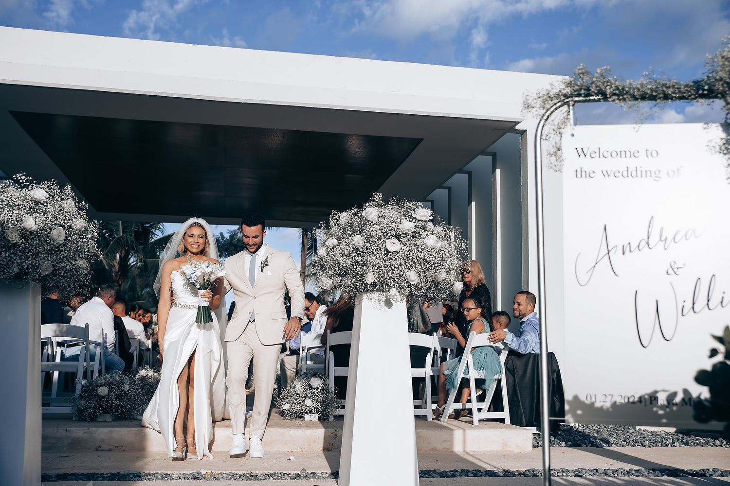 Couple exits tropical wedding ceremony venue under modern white architectural overhang with palm trees.