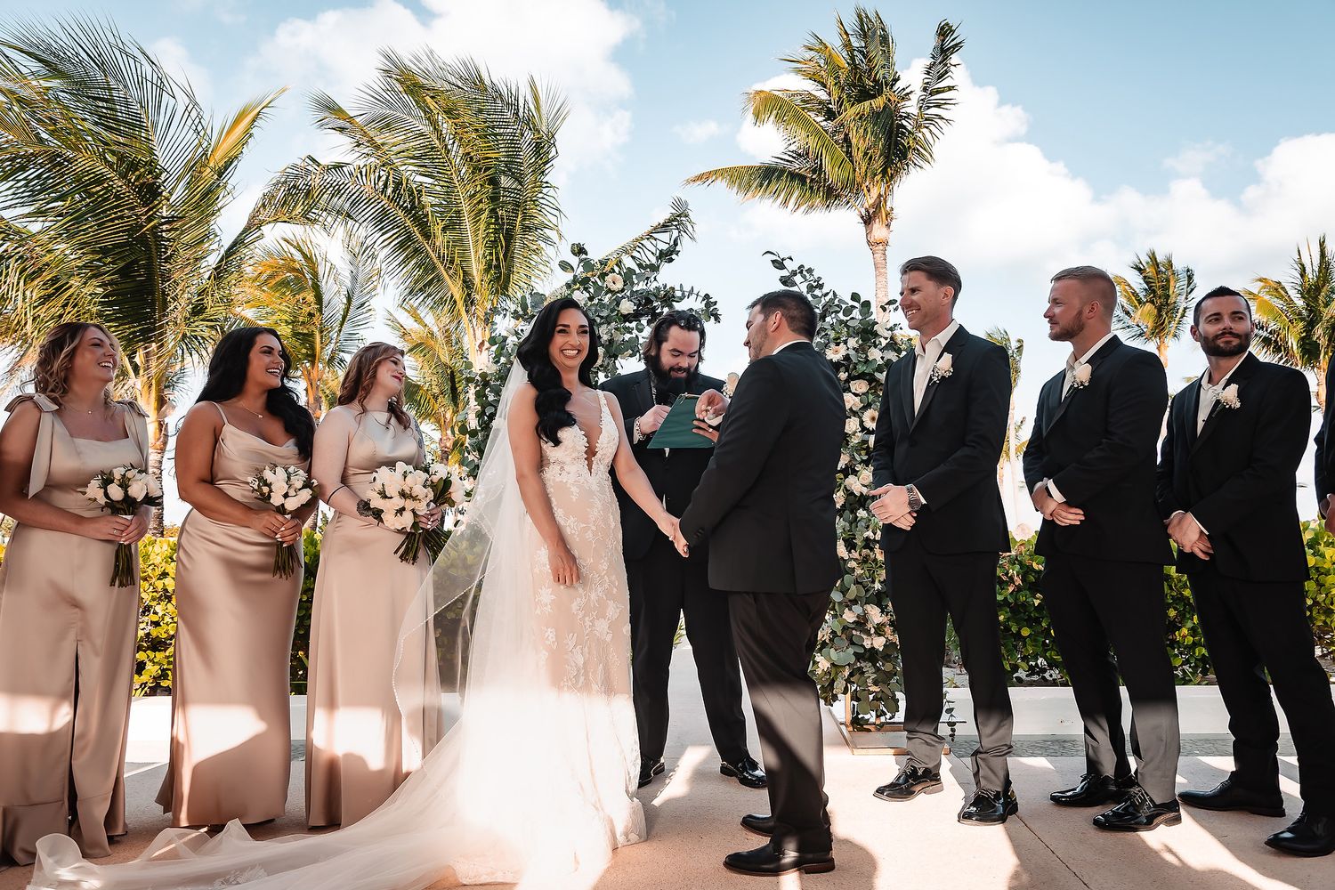 Outdoor wedding ceremony with bridesmaids in blush dresses and groomsmen in black suits.