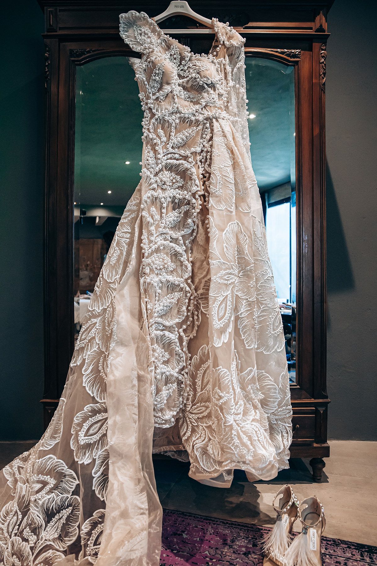 A delicate lace wedding dress with intricate embroidery details displayed on hangers against a dark background.