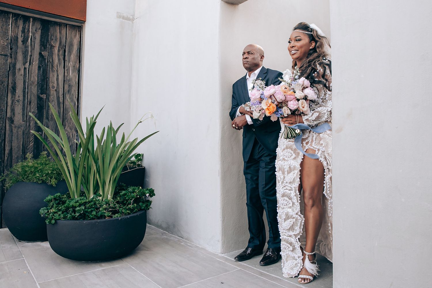 A couple stands against a white wall at a wedding ceremony, with the bride wearing a lace gown and holding a bouquet.
