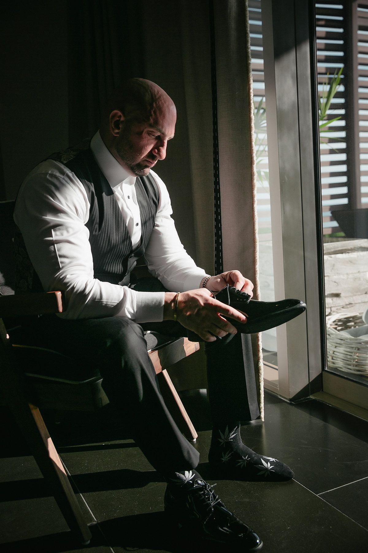 A man in formal attire sits by a window polishing a black dress shoe in moody lighting.