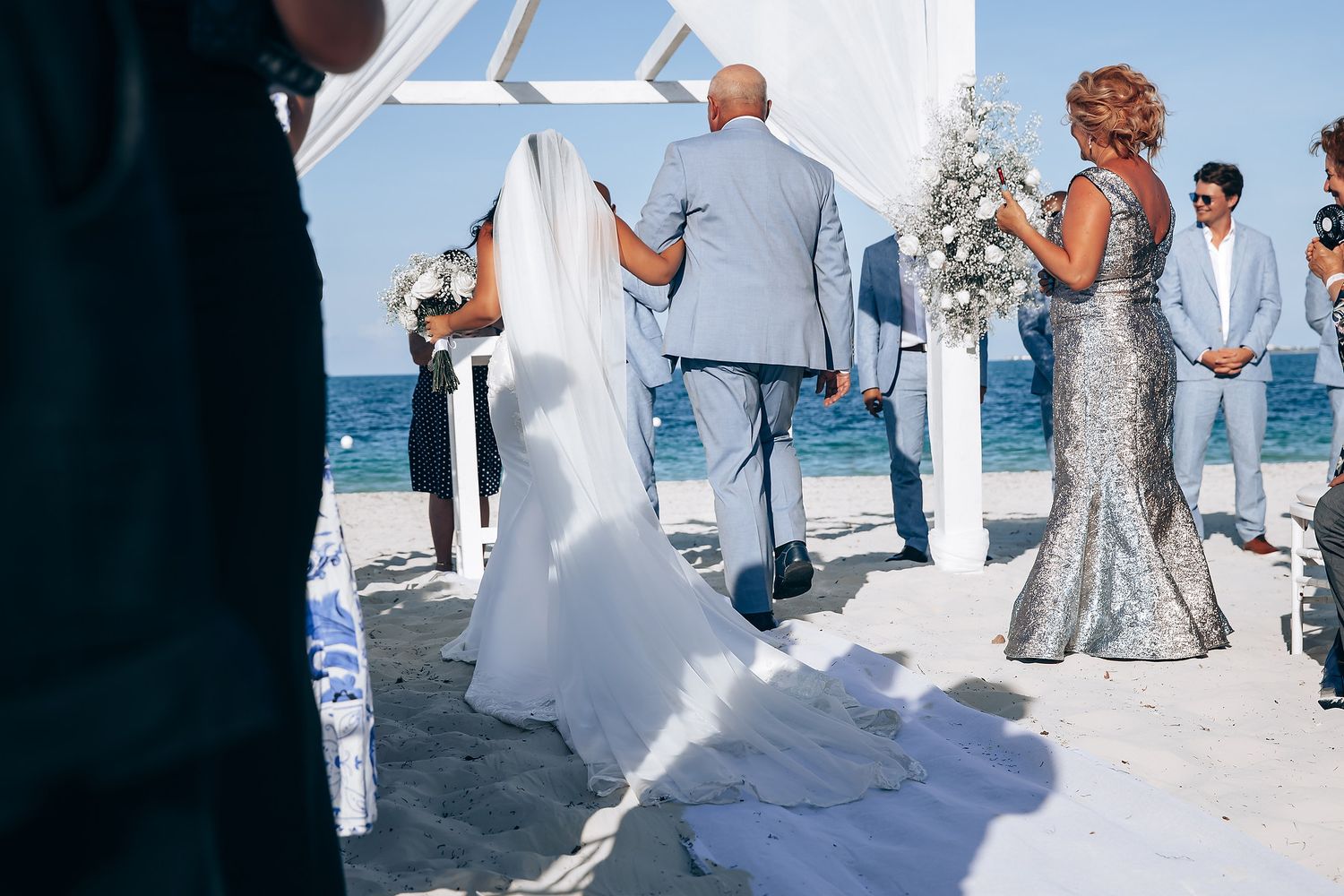 Elegant beach wedding ceremony with guests in light attire gathered on a sunny shoreline.