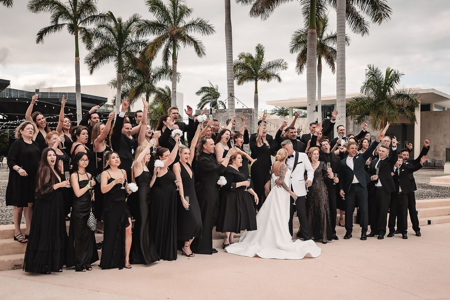 Wedding guests in black attire gather under palm trees for an outdoor celebration.