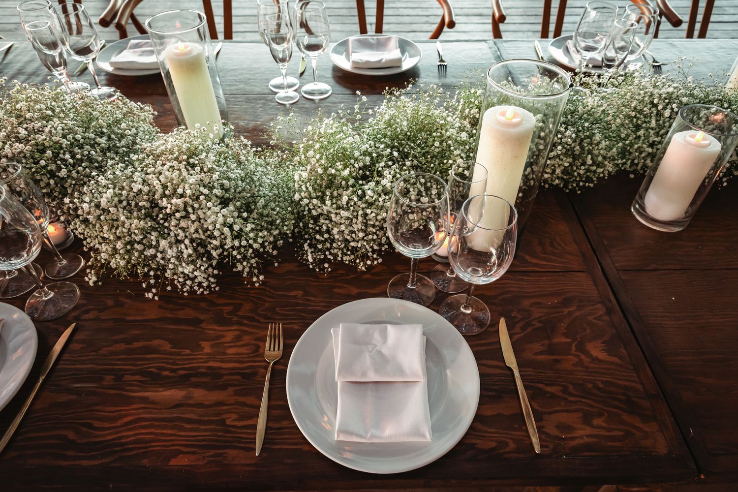 Elegant wedding reception table with baby's breath flowers, gold cutlery, candles, and gray linen napkins.