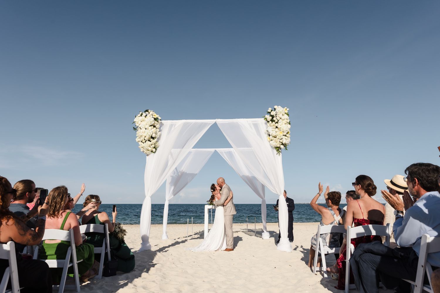 Couple exchanging vows at a beautiful beach wedding ceremony under a white floral arch with ocean backdrop.