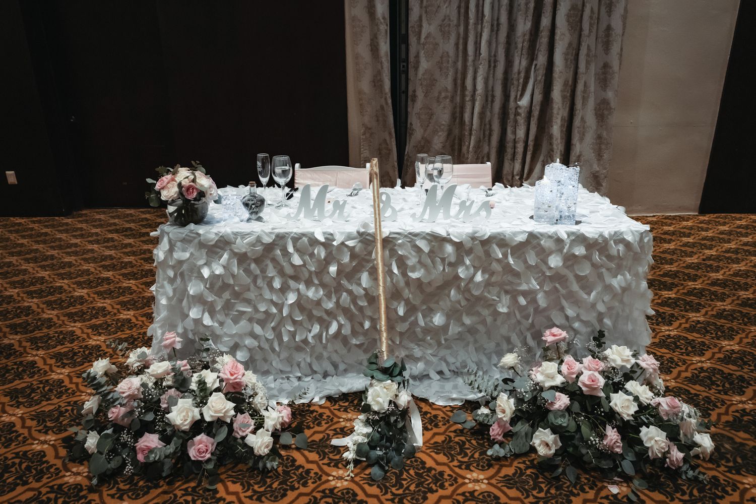 Elegant wedding sweetheart table with white textured linen, floral arrangements, and Mr. & Mrs. signs in a banquet hall.