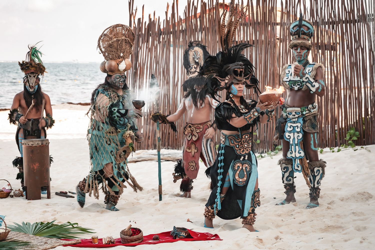 Four performers in traditional Mayan ceremonial costumes and headdresses performing a ritual dance on a sandy beach.