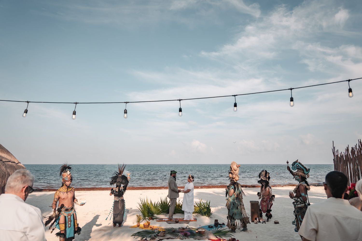 Traditional Mayan ceremony on a beach with performers in colorful costumes under string lights by the ocean.
