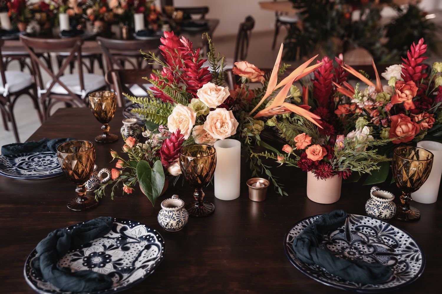Dark floral wedding tablescape with black patterned plates, lush burgundy and peach centerpieces, and amber glassware on a wooden table.
