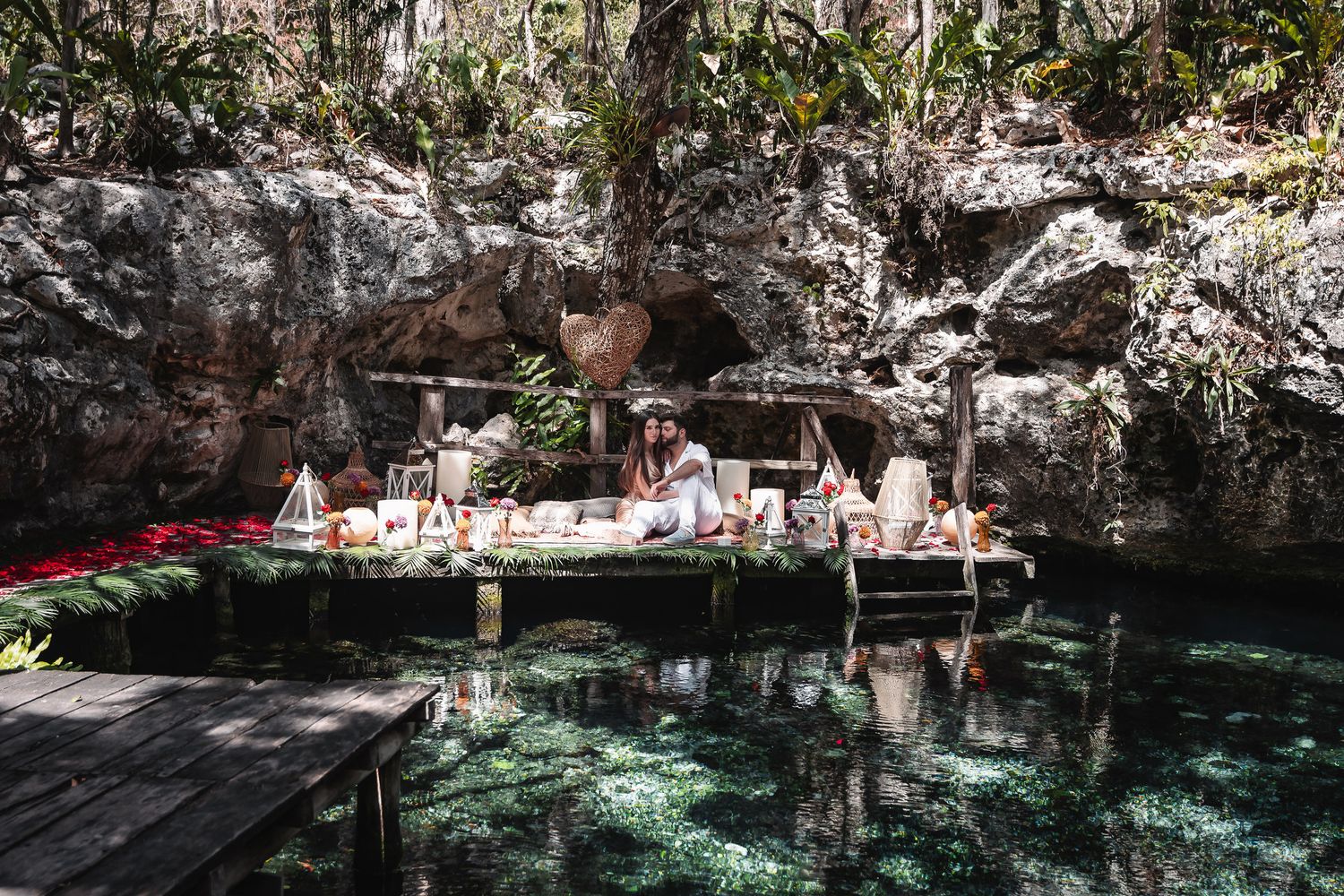 Woman sitting at a decorated table on a wooden dock beside a crystal-clear cenote surrounded by lush jungle rocks.