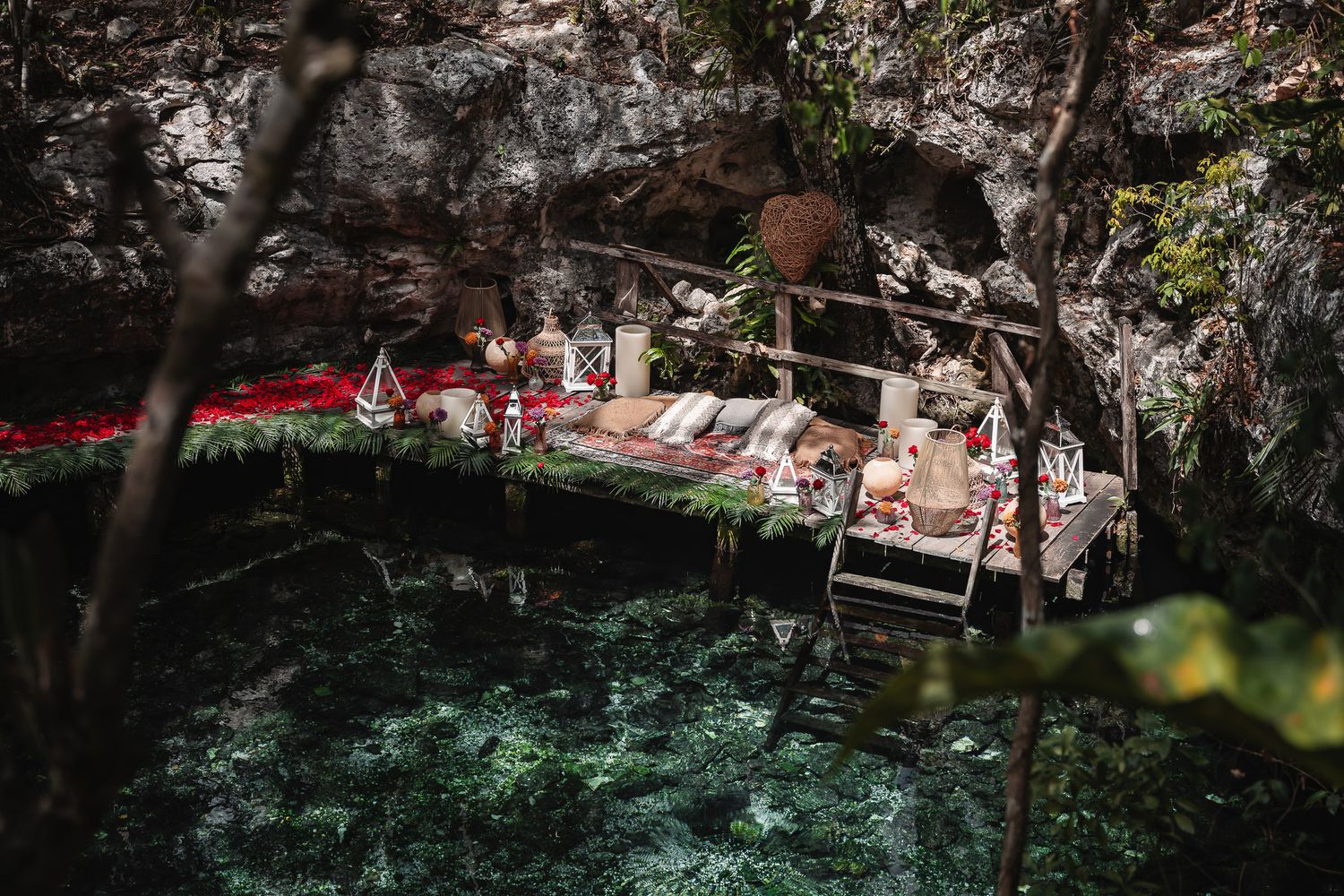 Romantic cenote picnic setup with red flowers, cushions, and wooden stairs over crystal-clear turquoise water in a cave.