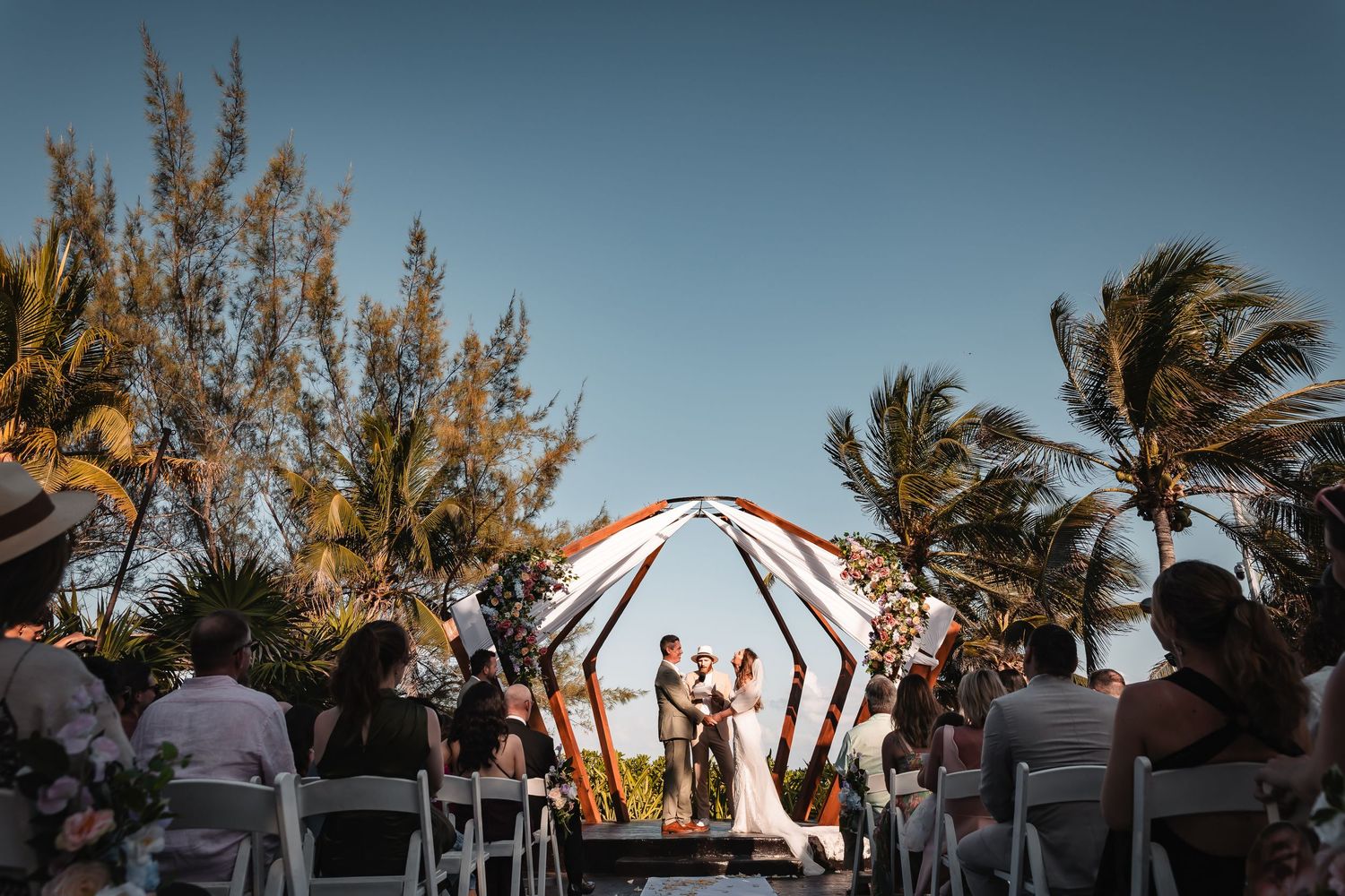 Wedding celebration under a canopy with palm trees silhouetted against a blue sky.