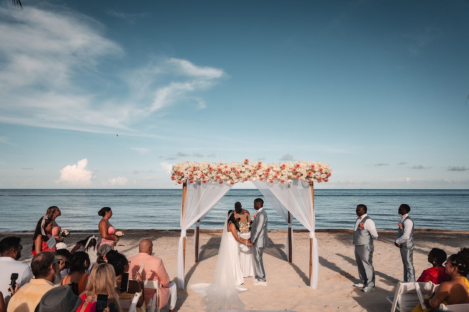 A panoramic view of a beach wedding ceremony with white chairs and floral arch against the ocean backdrop.