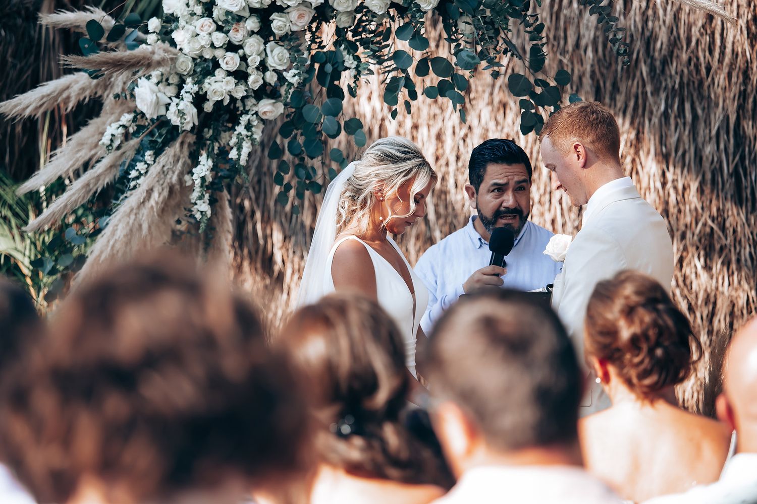 Outdoor wedding ceremony with guests seated beneath palm trees and white floral arrangements.