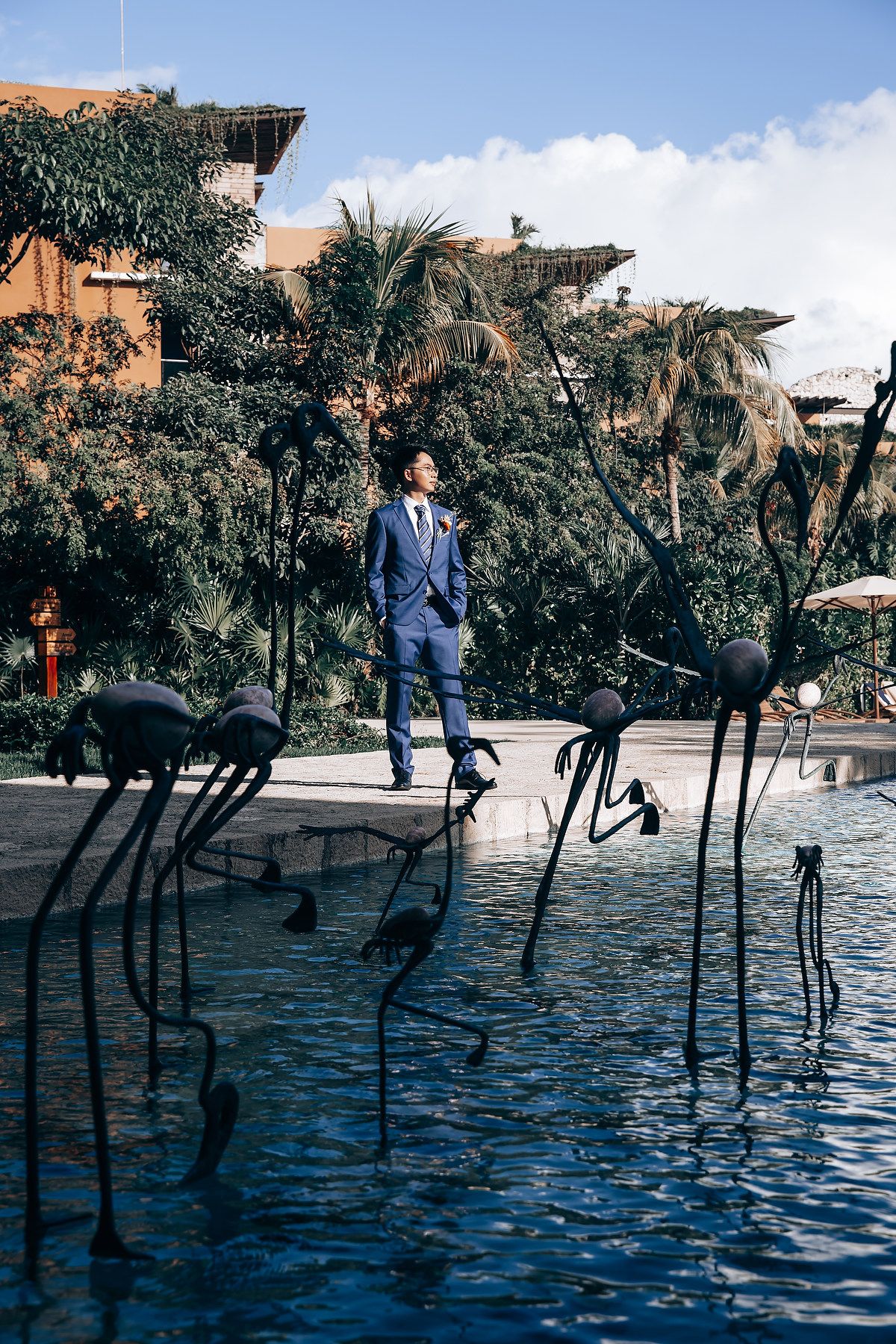 Blue-suited figure posed among dark metal sculptures in a reflective pool with palm trees.