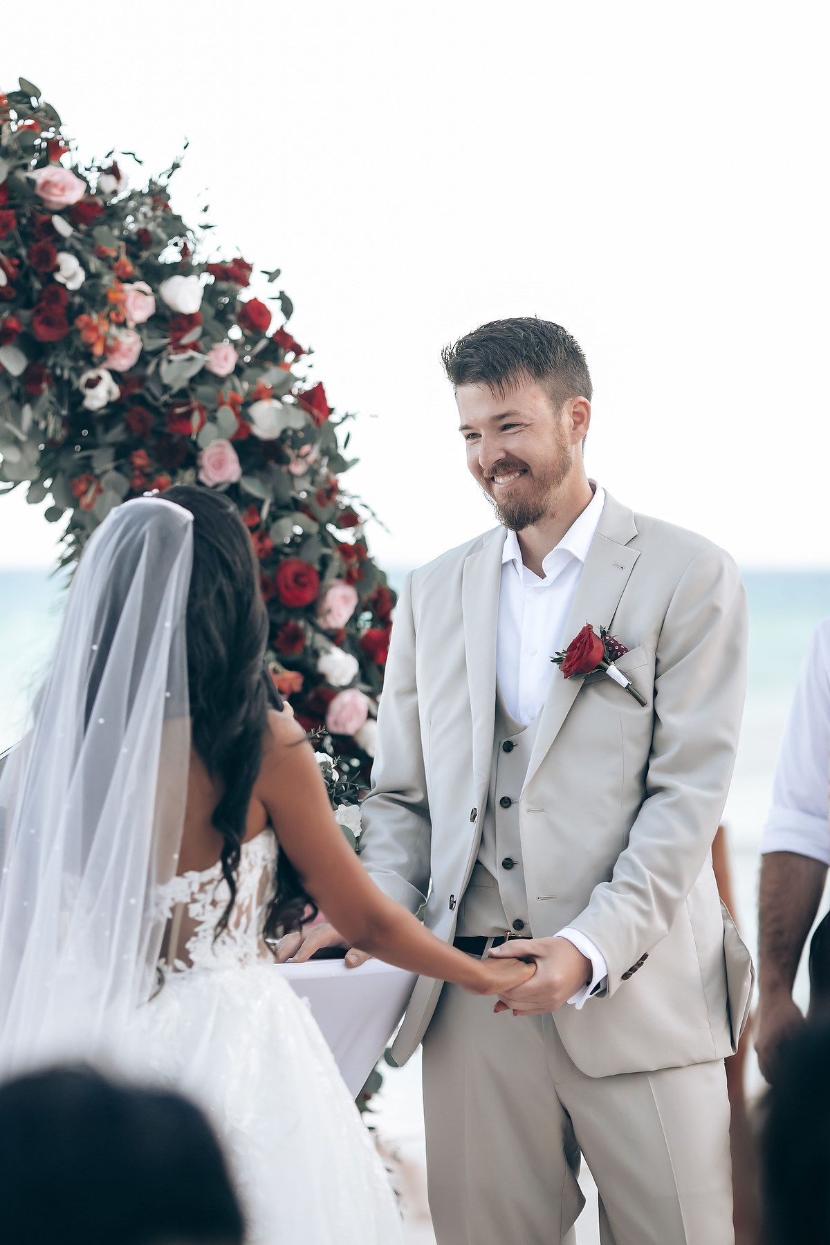 Wedding ceremony under floral arch with red roses against ocean backdrop.