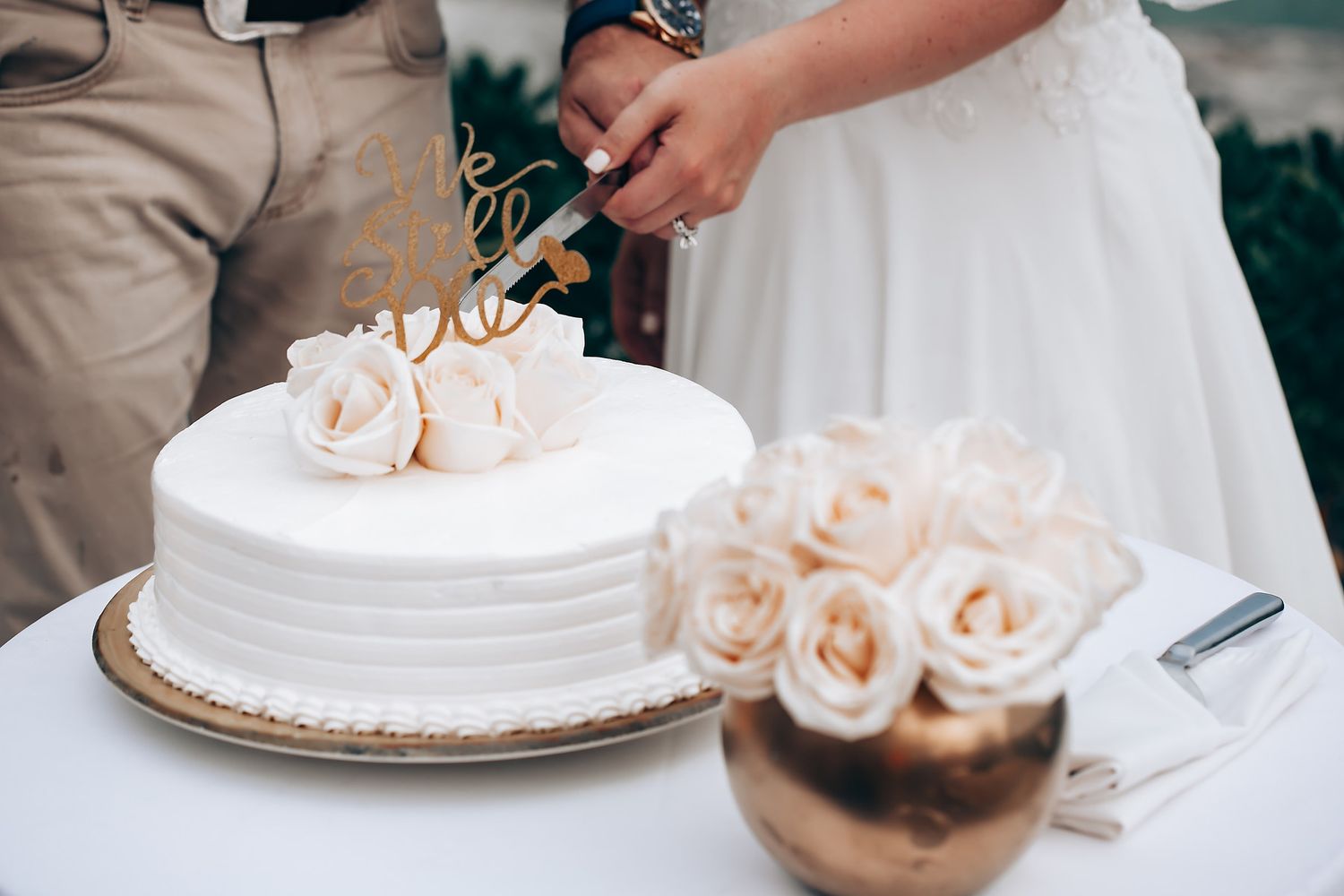 A white wedding cake decorated with blush roses sits on a table as someone cuts into it at a wedding reception.
