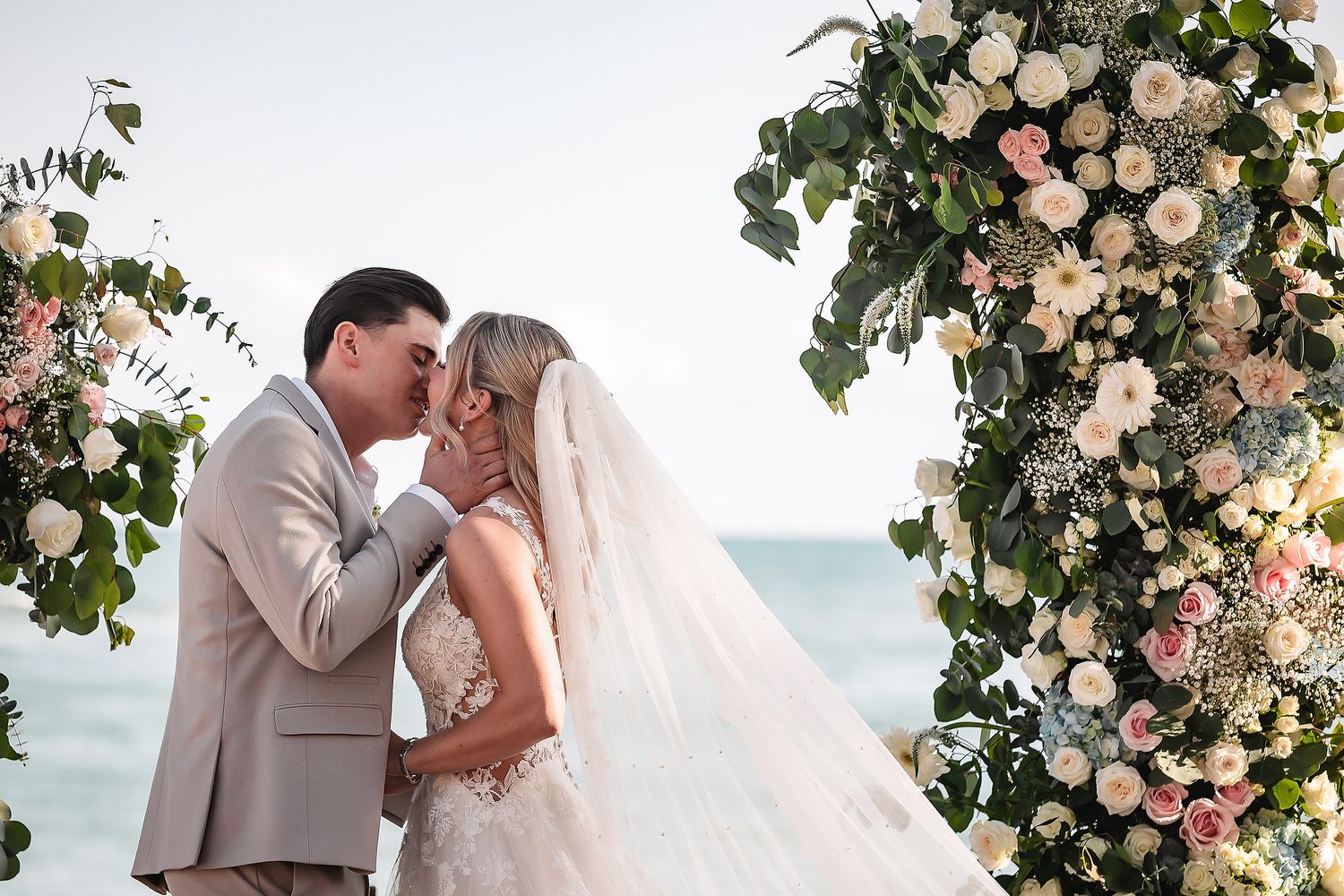 Wedding couple shares romantic moment under ornate floral arch with blush and white roses.