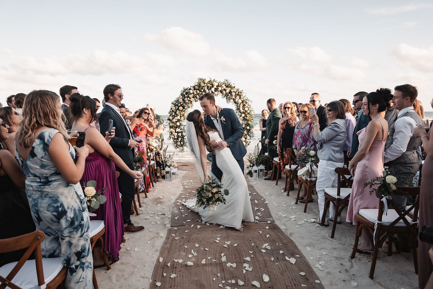 Wedding guests seated in rows at an outdoor ceremony with floral arch and scattered white petals.