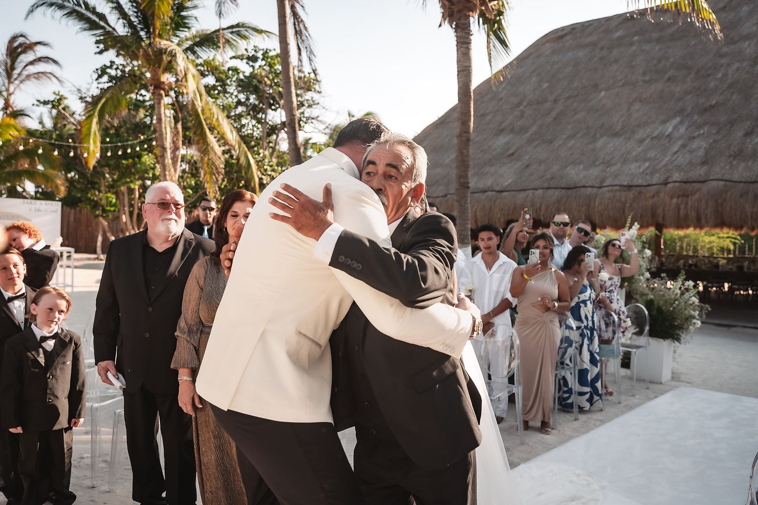 Joyful embrace during outdoor beach wedding ceremony with palm trees and guests looking on.