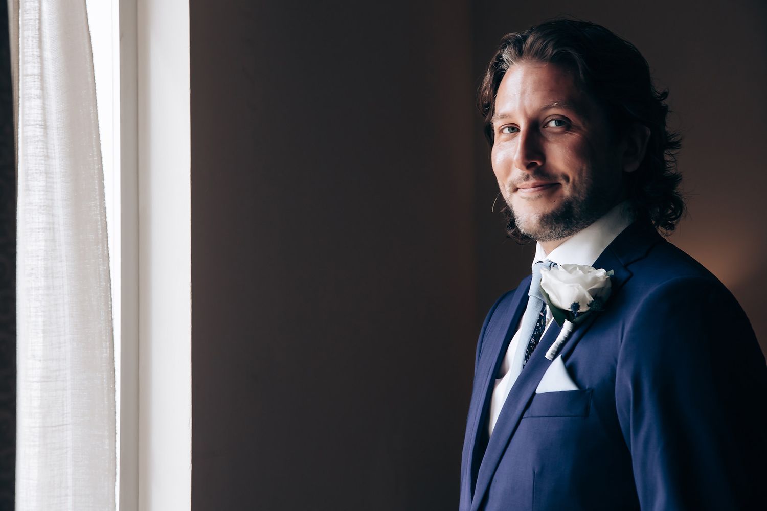 Professional portrait photo in dark navy suit standing by window with dramatic side lighting and moody atmosphere.