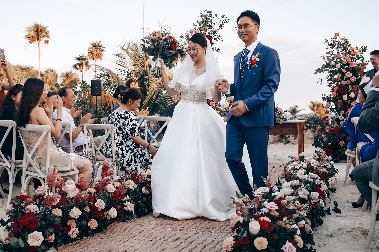 An outdoor wedding ceremony setup with pink and white floral arrangements lining the aisle at sunset.