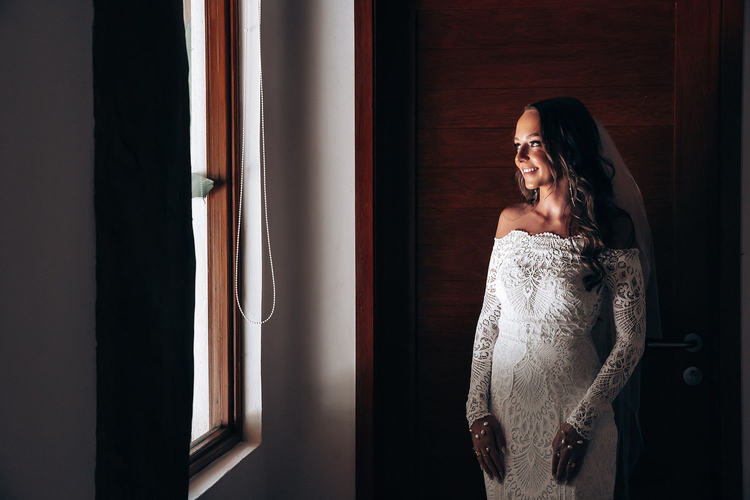 A person in a white lace dress stands near a window in dramatic lighting creating a moody silhouette.