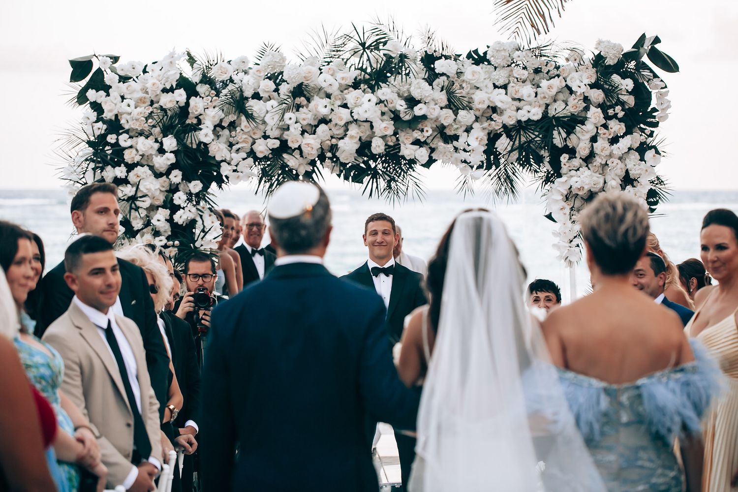 Wedding ceremony under a luxurious white floral arch overlooking the ocean.