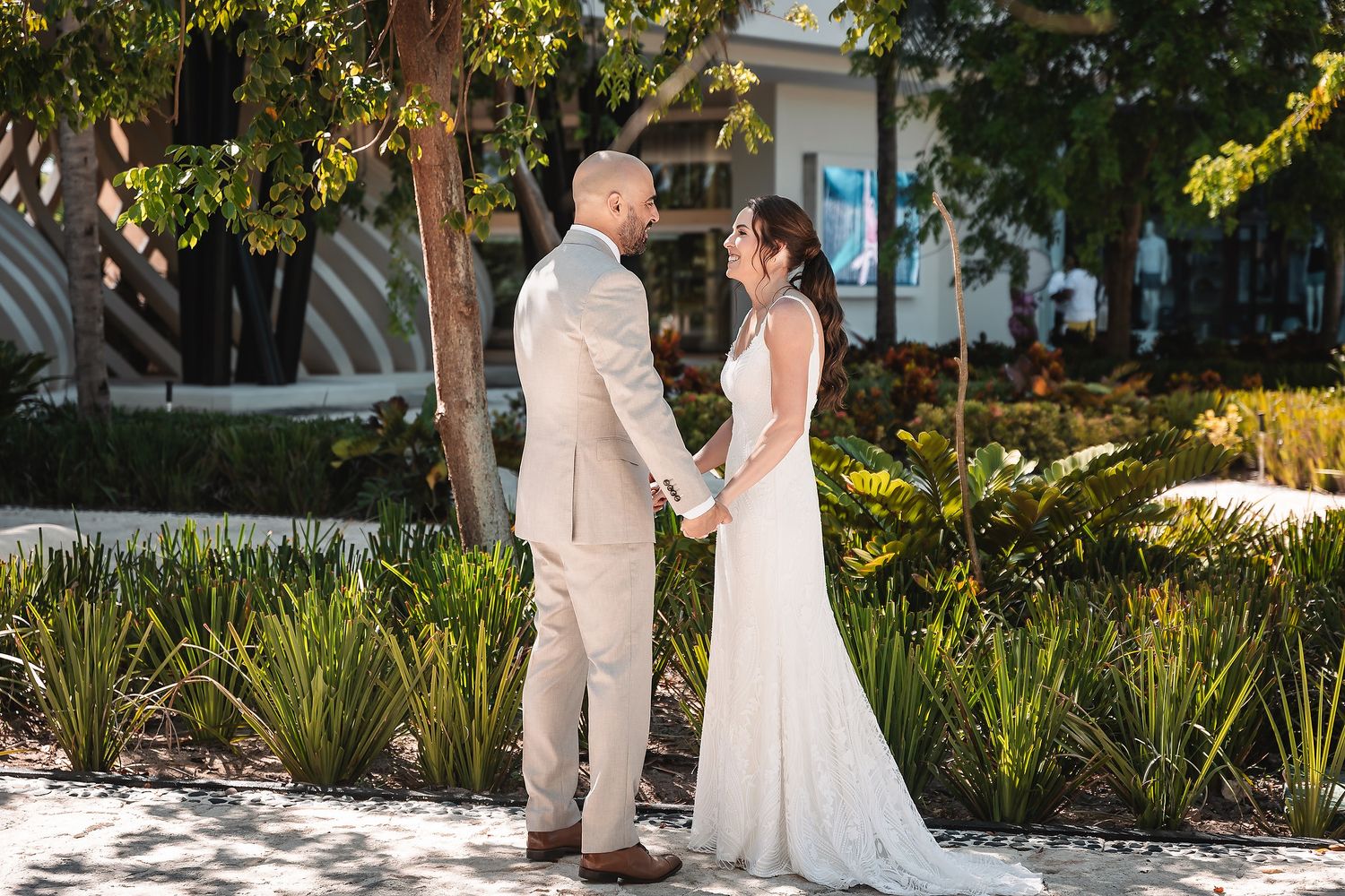 Two people in wedding attire share an intimate moment in a lush garden setting during a sunny day.