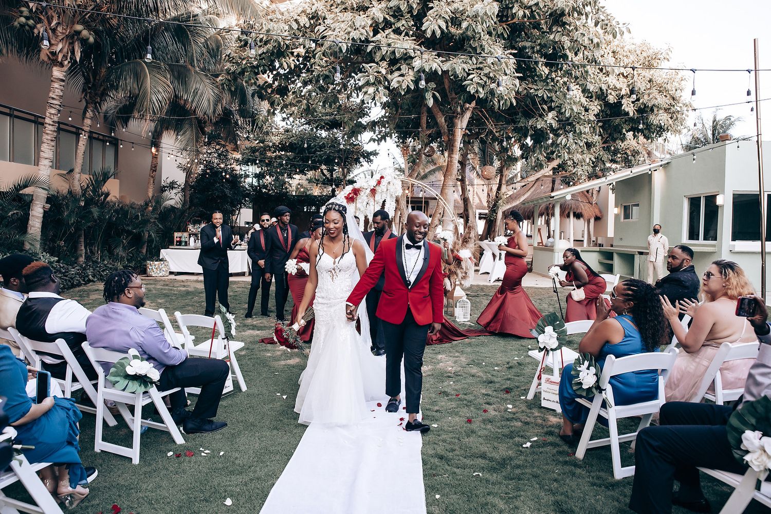 Wedding guests gather outdoors on white chairs during a garden ceremony with trees and buildings in the background.