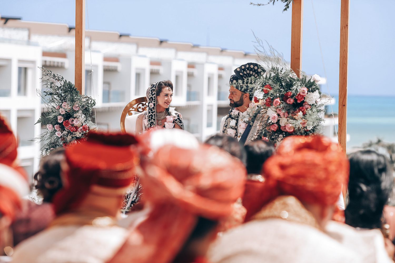 Traditional Balinese wedding ceremony at a beachfront venue with decorative red headdresses in the foreground.