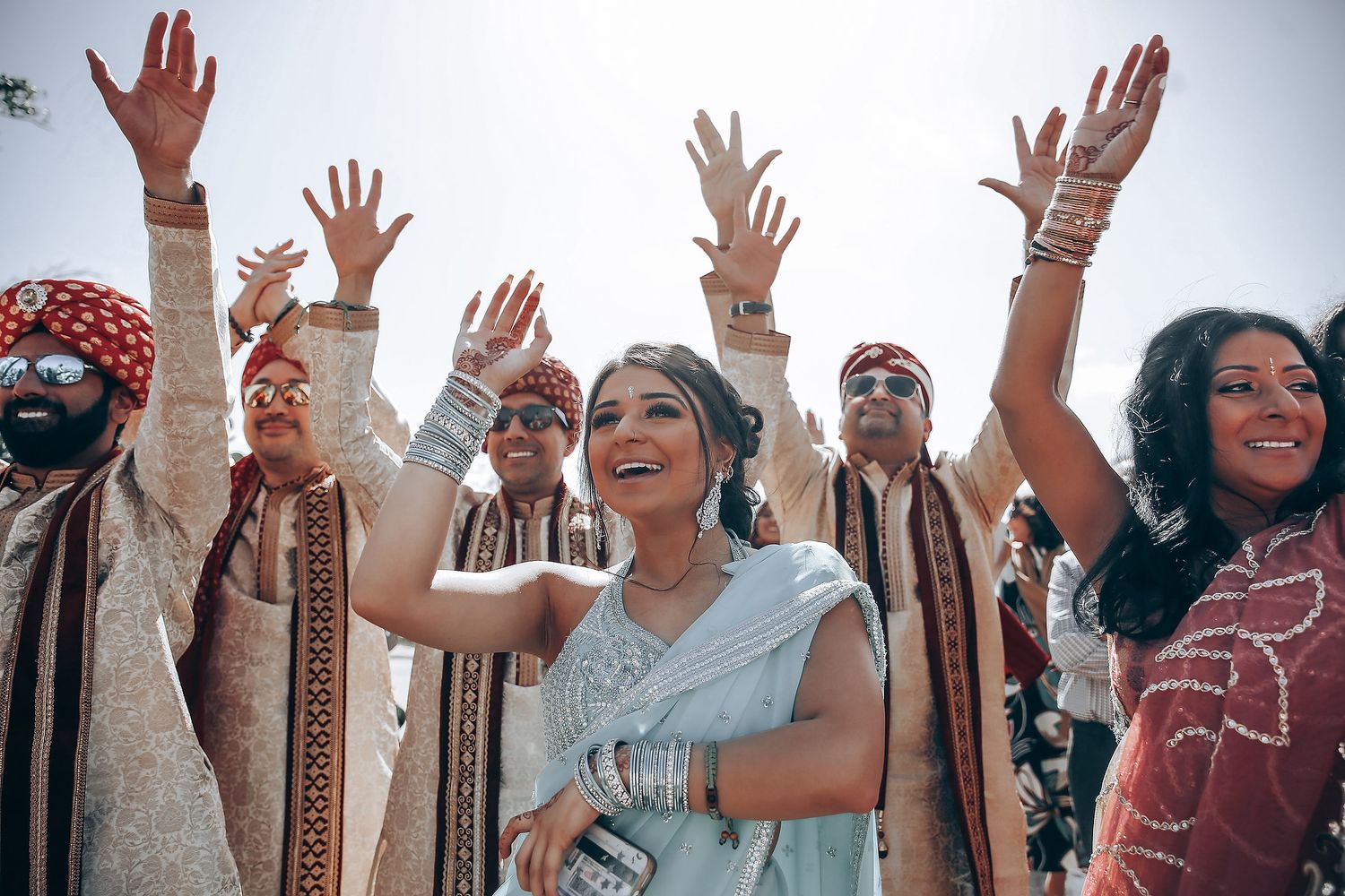 Joyful wedding celebration with people raising their hands in traditional Indian attire during outdoor ceremony.
