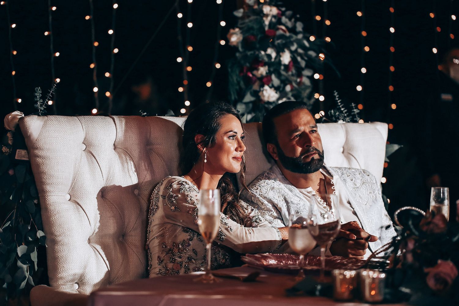 A couple enjoys an intimate dinner setting with tufted seating and twinkling lights in the background.