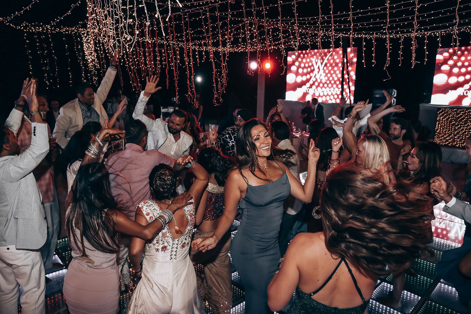 A crowded nightclub scene with people dancing and socializing under red lighting and decorative string lights.