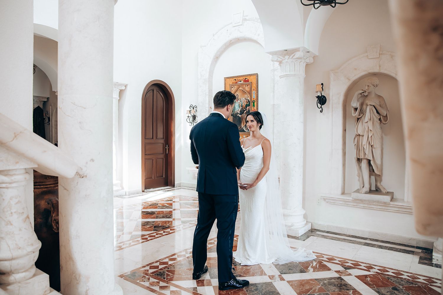 A couple shares an intimate moment in an elegant white marble hallway with arched doorways and classical statues.