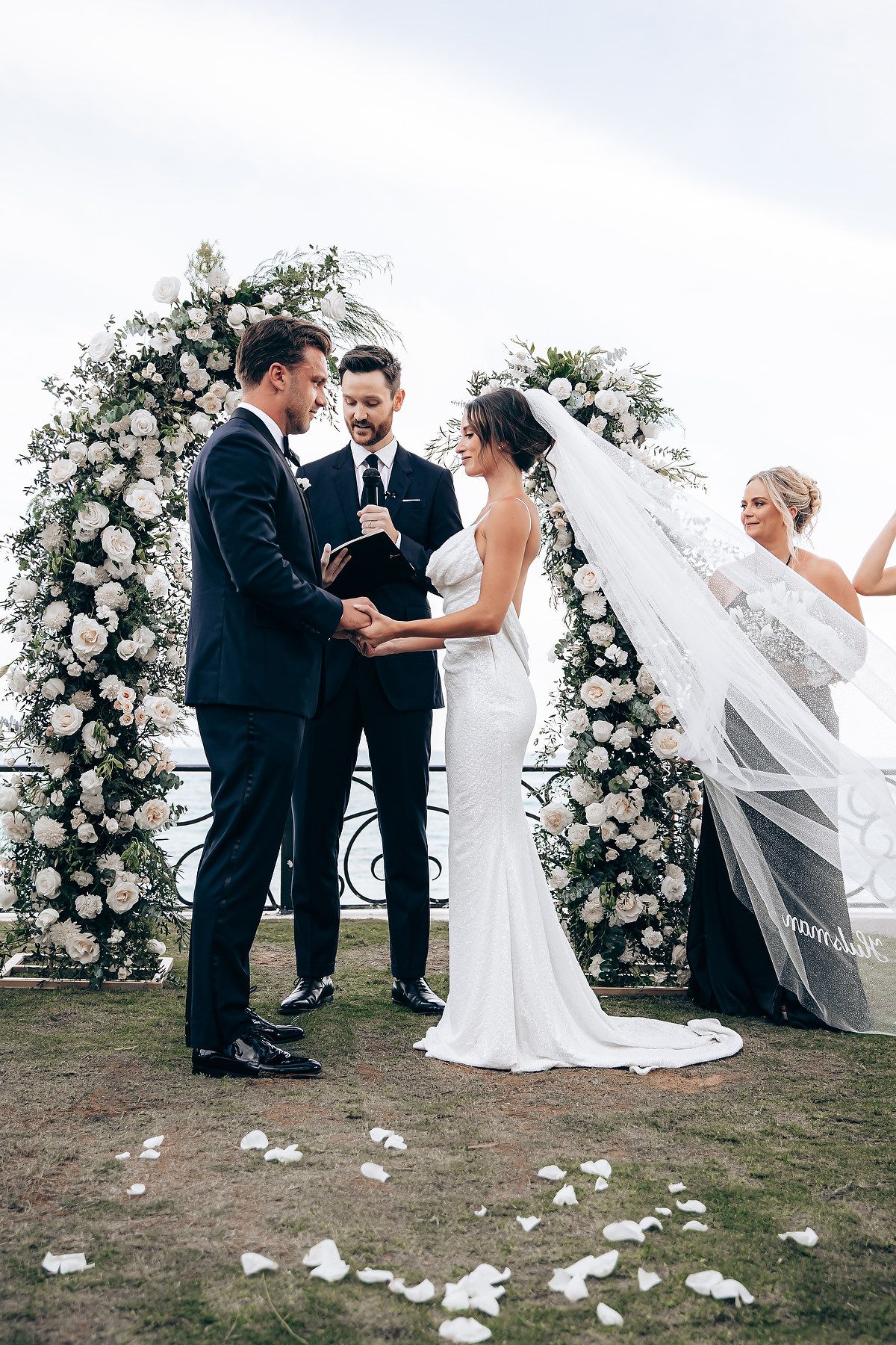 A wedding ceremony takes place under a floral arch with white roses as a veil catches the breeze.