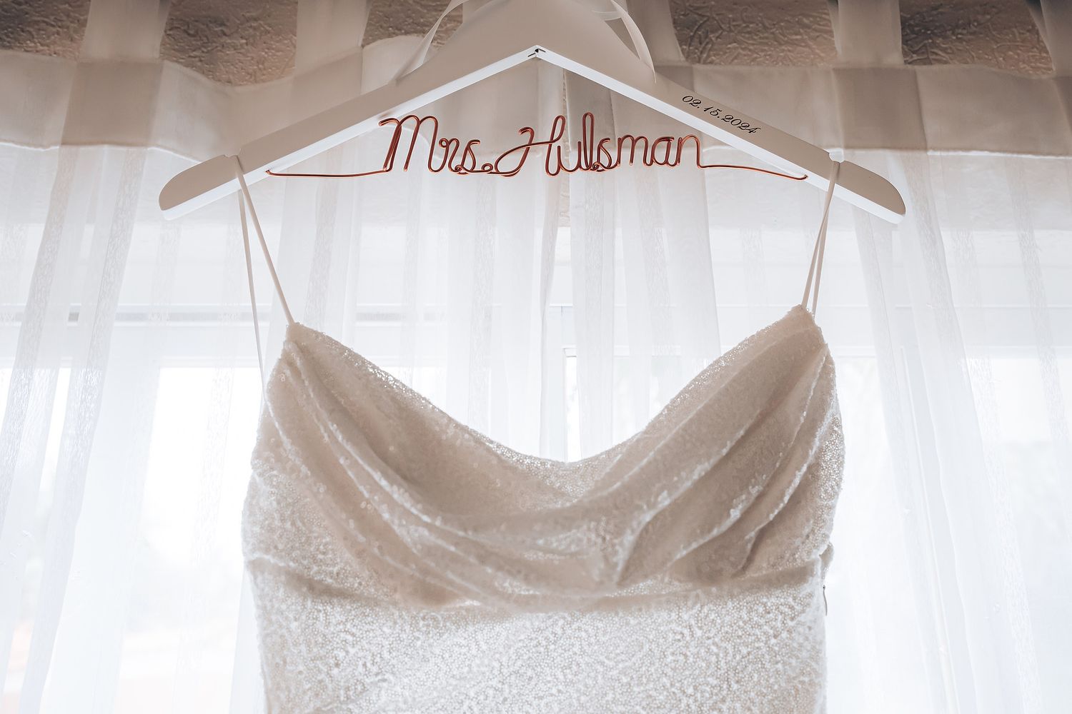 White wedding dress hanging on a personalized hanger against a light background with sheer curtains.