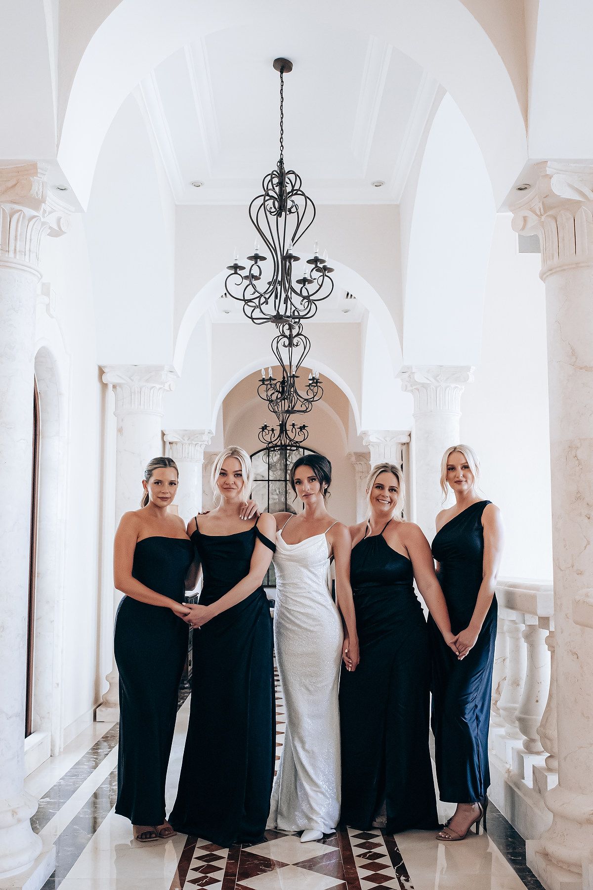 Elegant bridal party poses in formal dresses under ornate chandeliers in a white columned hallway.