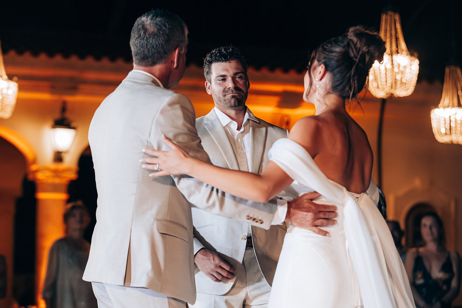 Wedding guests dressed in white dance and embrace on a warmly lit dance floor during an evening celebration.