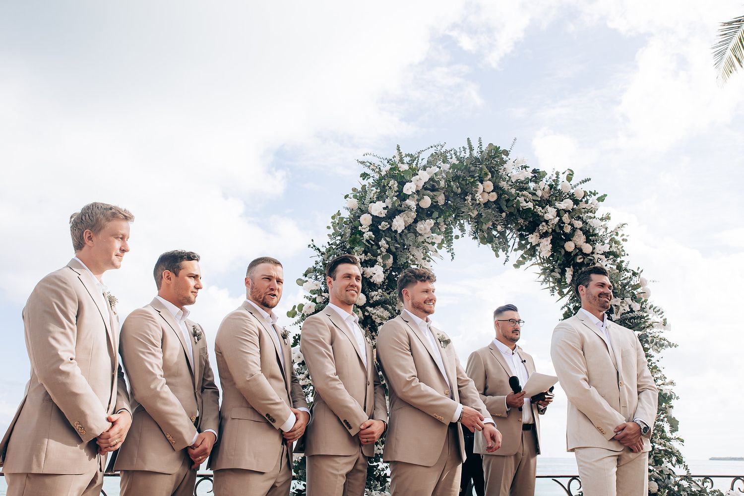 Wedding party in tan suits stands in a line on a grassy hill against a bright sky background.