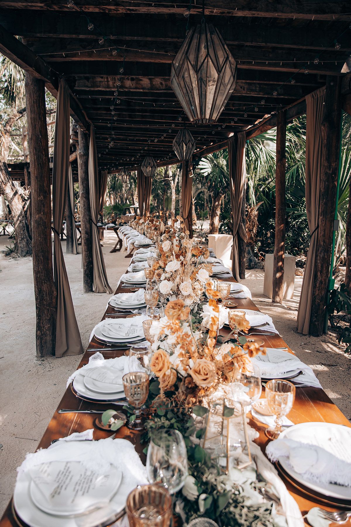 Elegant rustic wedding reception table setting with dried flowers, lanterns and wooden beams in an outdoor covered venue.
