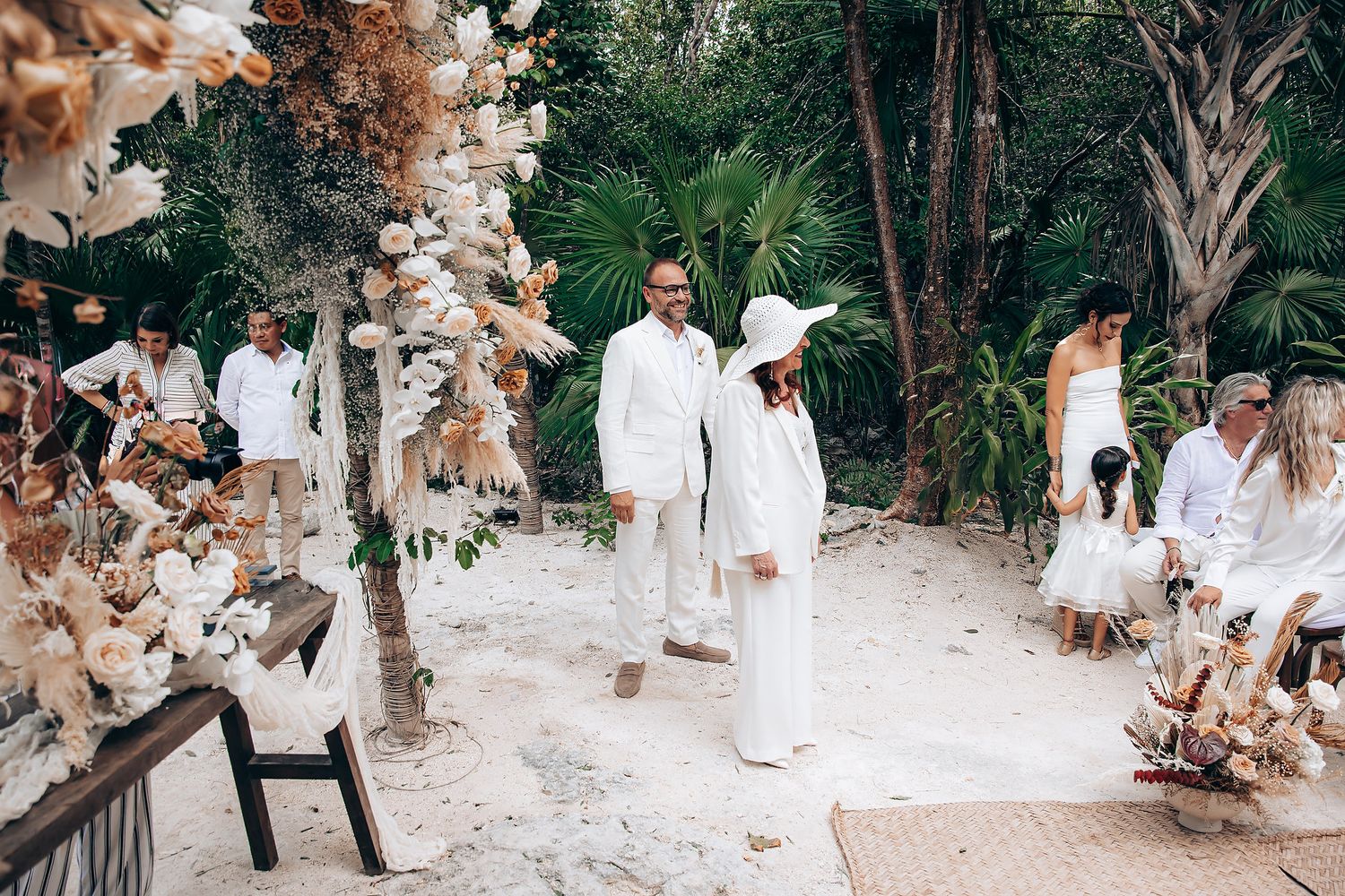 Wedding guests in white attire gather outdoors under trees during a tropical beach ceremony.
