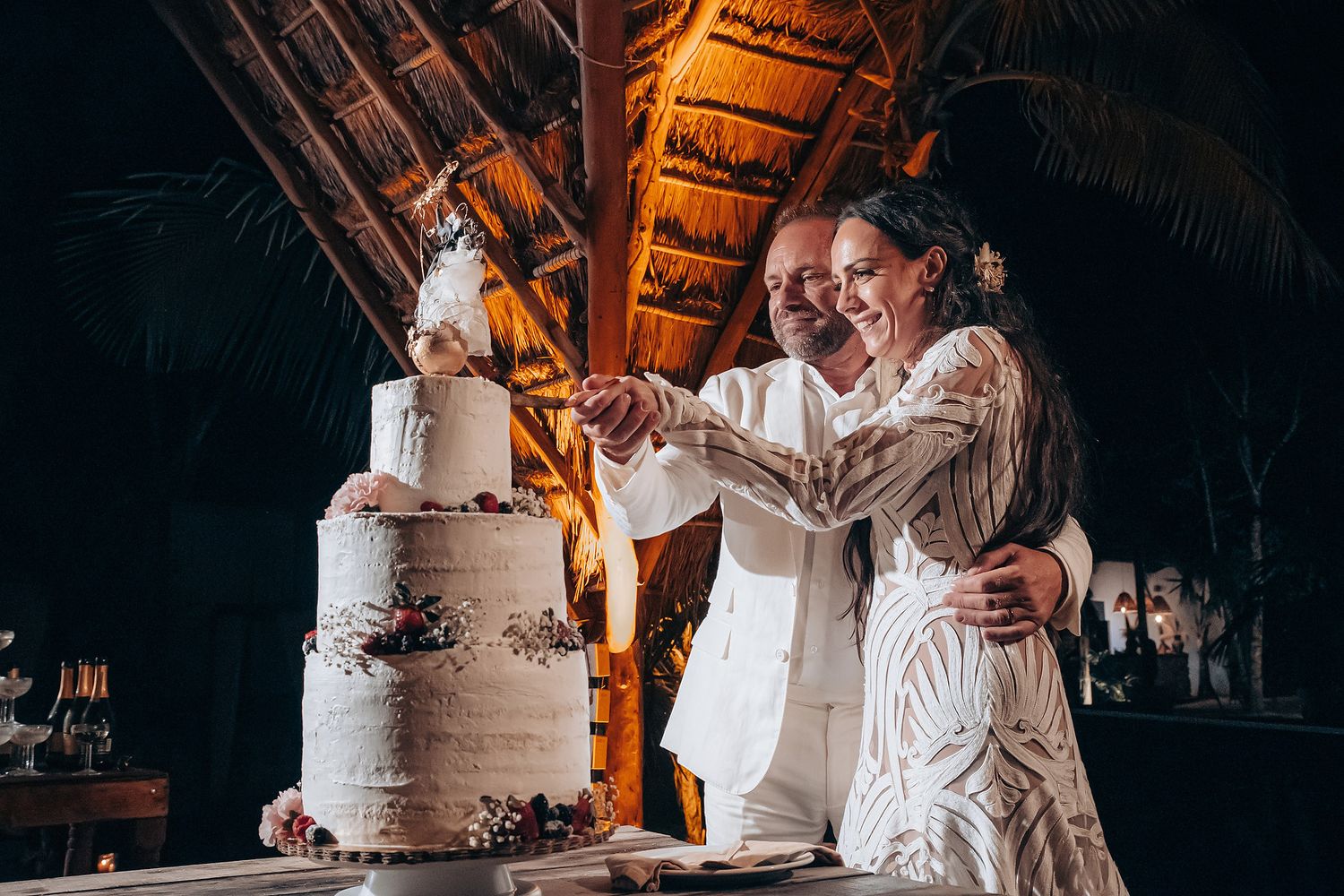 A wedding couple shares a romantic moment while cutting their white tiered cake in warm ambient lighting.