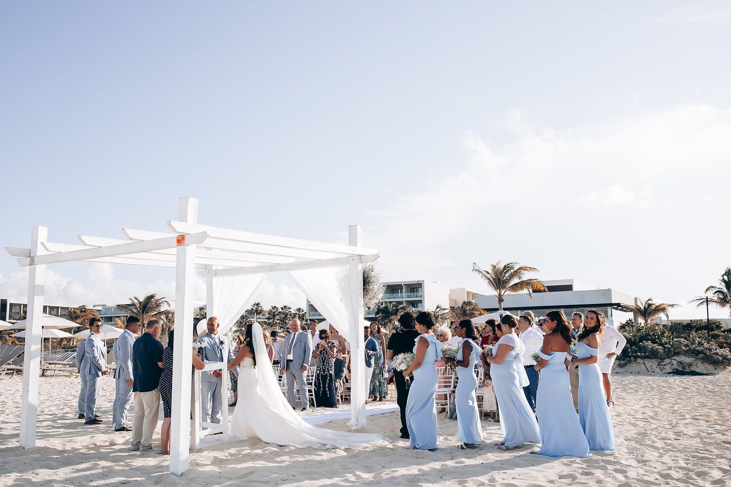 Panoramic view of a beach wedding ceremony with white decor and guests seated on white chairs.