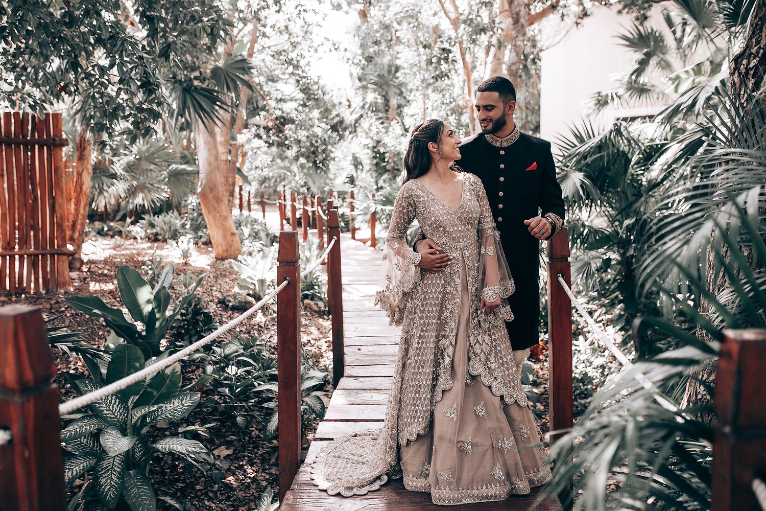 Couple walking along wooden pathway through tropical garden during wedding photoshoot.