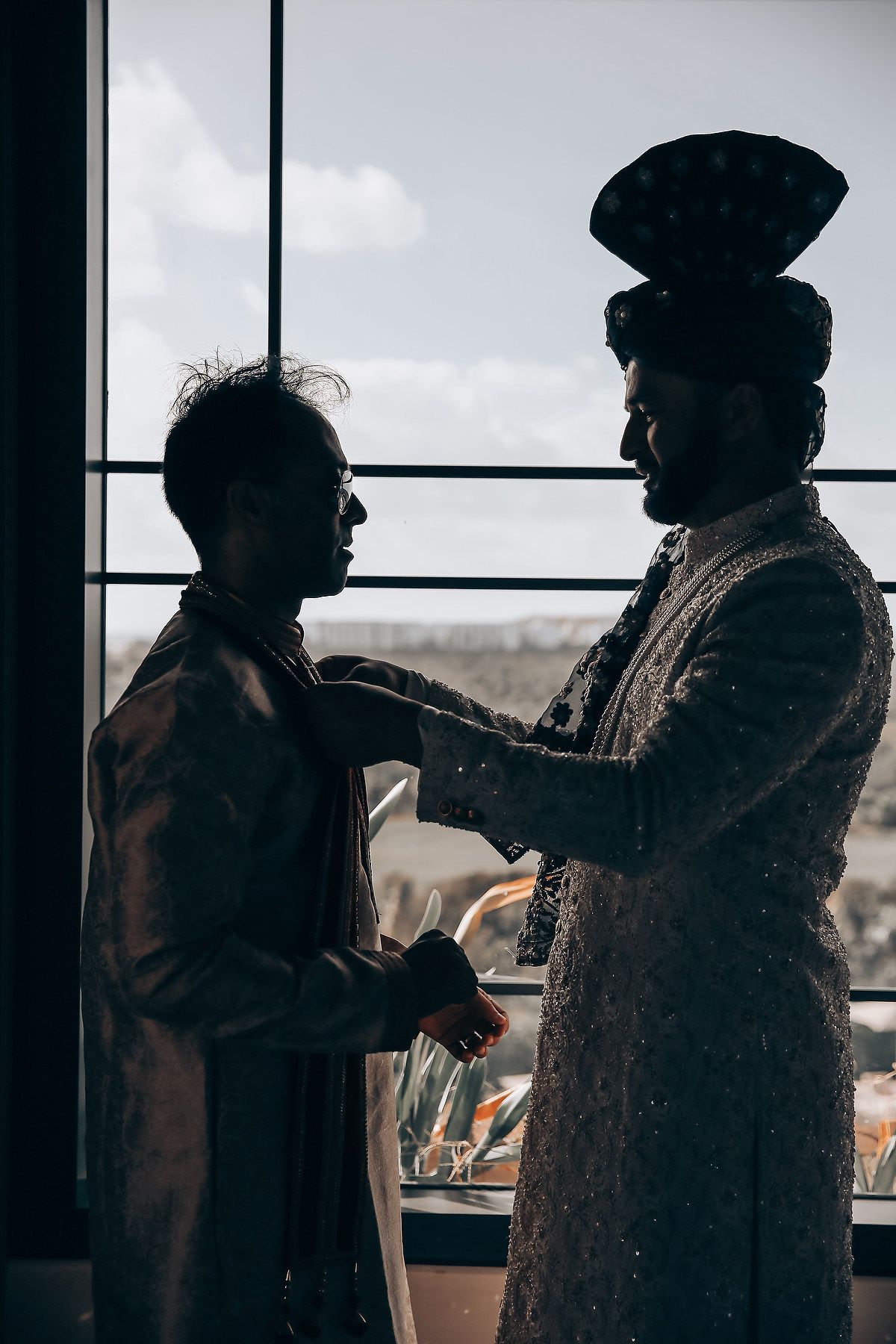 Silhouettes of people getting ready in traditional Indian wedding attire against a bright window.