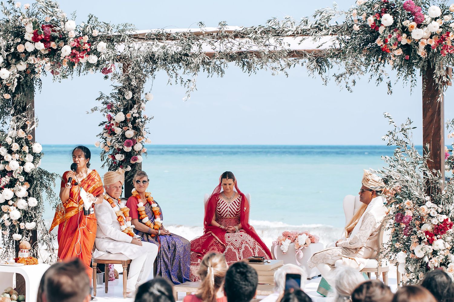 Beachfront wedding ceremony with guests seated under a floral-decorated wooden arch by the ocean.